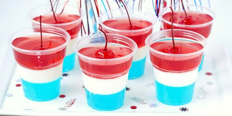 Close up of a collection of blue, white and red layered jello shots in plastic shot glasses, garnished with red cherries