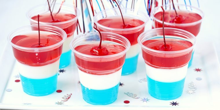 Close up of a collection of blue, white and red layered jello shots in plastic shot glasses, garnished with red cherries