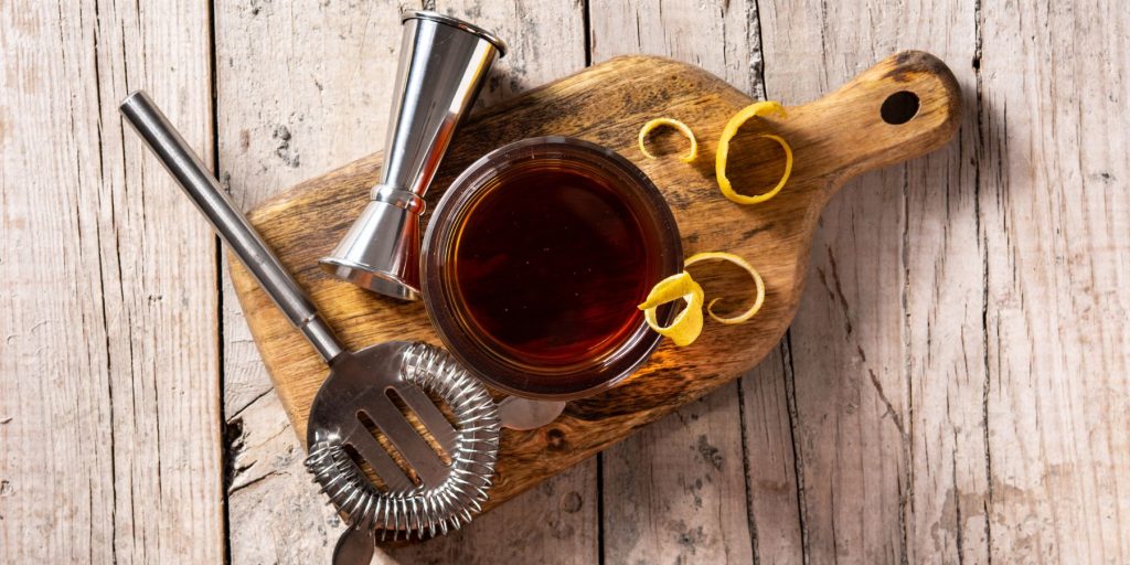 A top view of a single Sazerac cocktail served on a wooden serving platter, along with a cocktail jigger and strainer