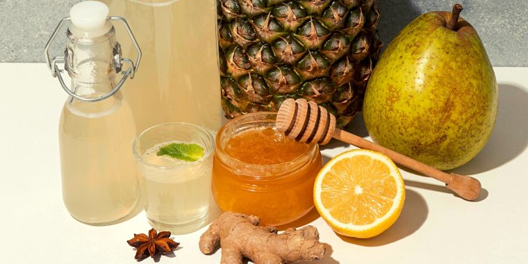 Selection of fermented cocktails and fruit on a white surface along with a jar of honey, a ginger root, halved lemon and star anise