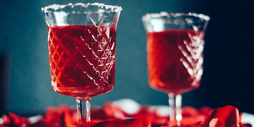 A beautiful pair of Love Potion #9 cocktails for Valentine's Day in tall, stemmed cut-glass tumblers, surrounded by rose petals against a blue backdrop