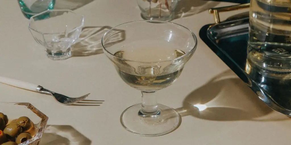 A simply elegant Irish Martini cocktail in a clear cocktail glass, arranged on a white tablecloth surrounded by cultery, a decanter and a bowl of green olives
