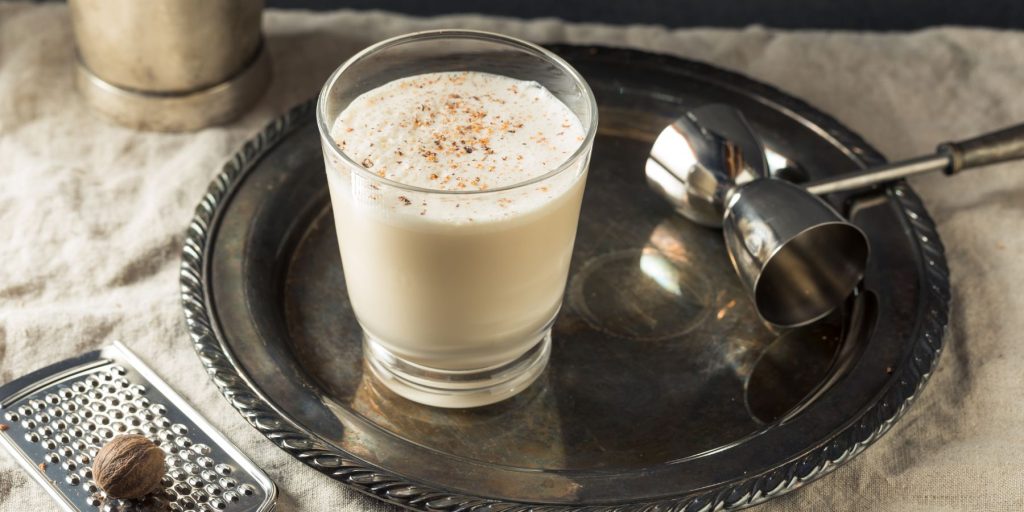 A tumbler of Brandy Milk Cocktial set on a silver serving platter on a table with a white tablecloth, flanked by a silver jigger and a silver microplane grater with a piece of whole nutmeg