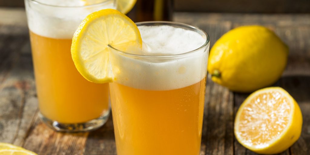 Two yellow-tinged Big Game Shandy cocktails on a wooden surface with sliced lemons in the background