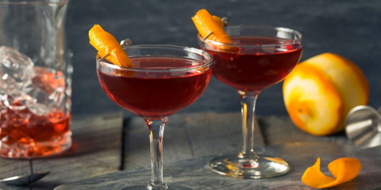 Two bourbon-based Revolver Cocktails garnished with flamed orange twists against a pewter backdrop with a peeled orange and a container of bourbon in the background
