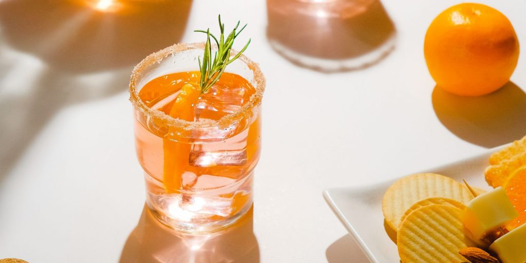 Close-up of an orange hued cocktail with a sugar rim and rosemary garnish on a white surface, surrounded by a clementine and a plate of snacks