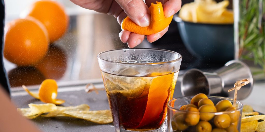 A close up of a home mixologist squeezing an orange twist into a refreshing cocktail with a bowl of olives in the foreground and fresh oranges in the background