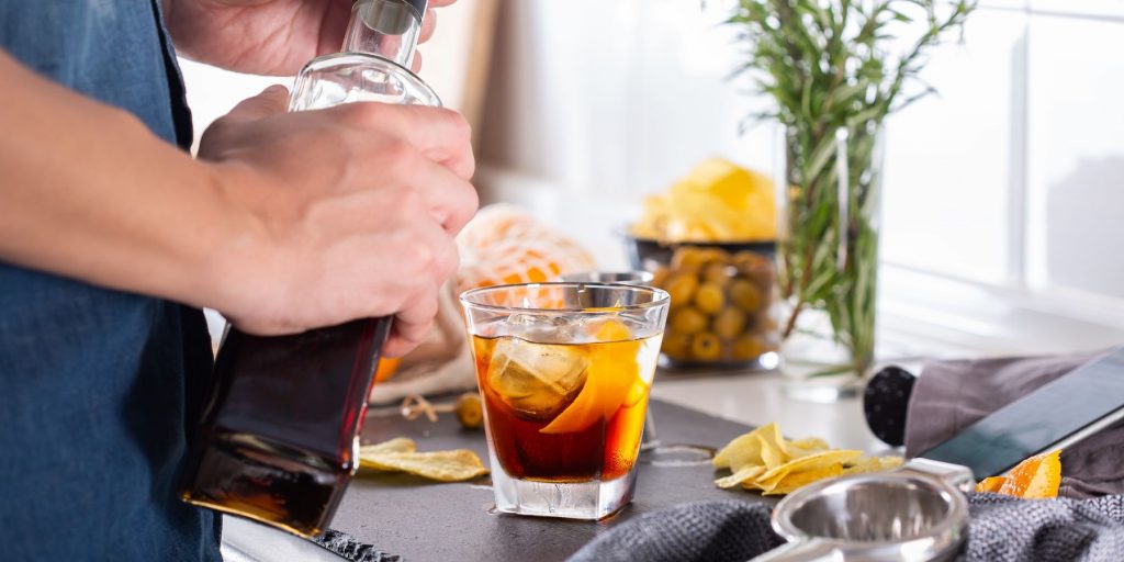 Side view of a home mixologist pouring holding a bottle of orange liqueur next to a surface filled with cocktail making tools and a refreshing cocktail garnished with an orange twist