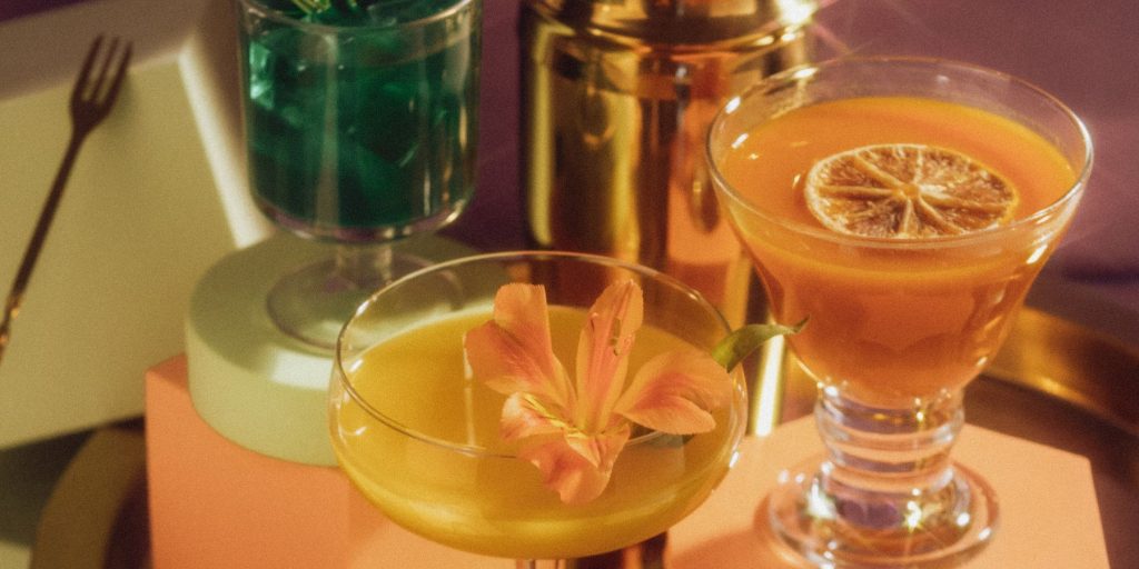 A close up of two retro-looking orange cocktails on a dusty pink podium with a golden cocktail shaker in the background