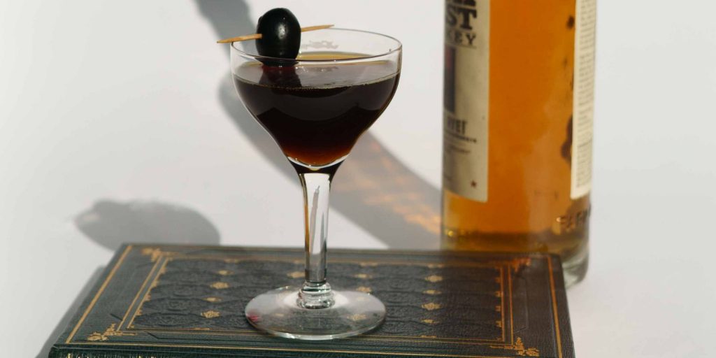 Close up of a dark-hued Bijou cocktailk in a coupe glass, garnished with a Luxrado cherry with a bottle of liqueur to the righthand side