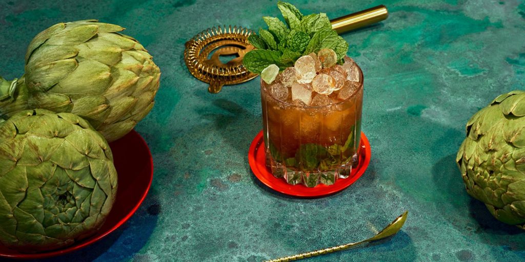 Close-up of a Cynar Julep on a blue marble surface surrounded by fresh artichokes
