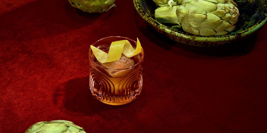 A close-up of a Bitter Guiseppe cocktail on a red velvet backdrop surrounded by fresh artichokes