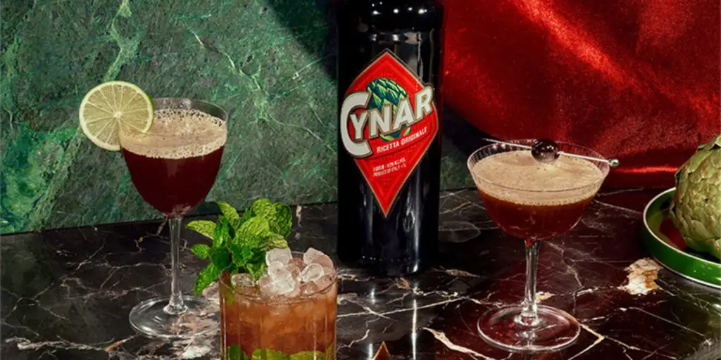 Close up of a bottle of Cynar surrounded by cocktails and fresh artichokes against a blue marble backdrop draped with red velvet