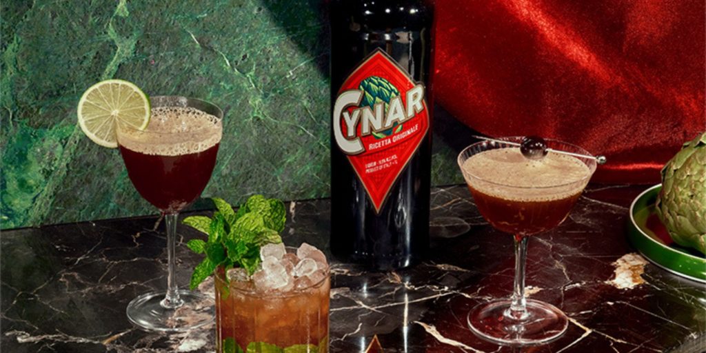 Close up of a bottle of Cynar surrounded by cocktails and fresh artichokes against a blue marble backdrop draped with red velvet