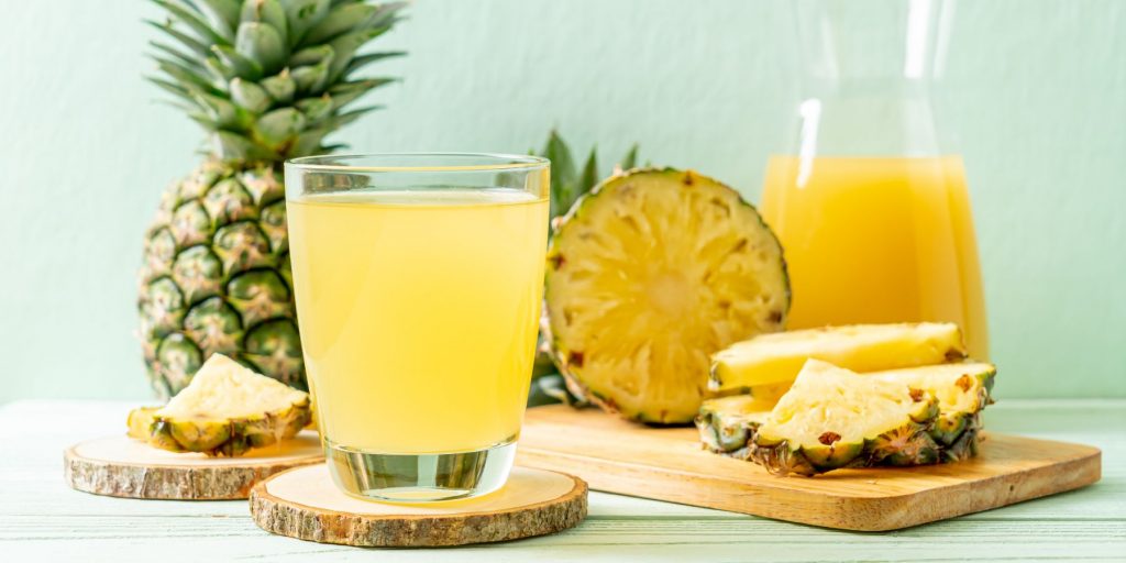Front view of a bubbly Pineapple Mimosa in a rocks glass on a wooden coaster with a jug of fresh pineapple juice and sliced pineapples in the background against a light blue backdrop