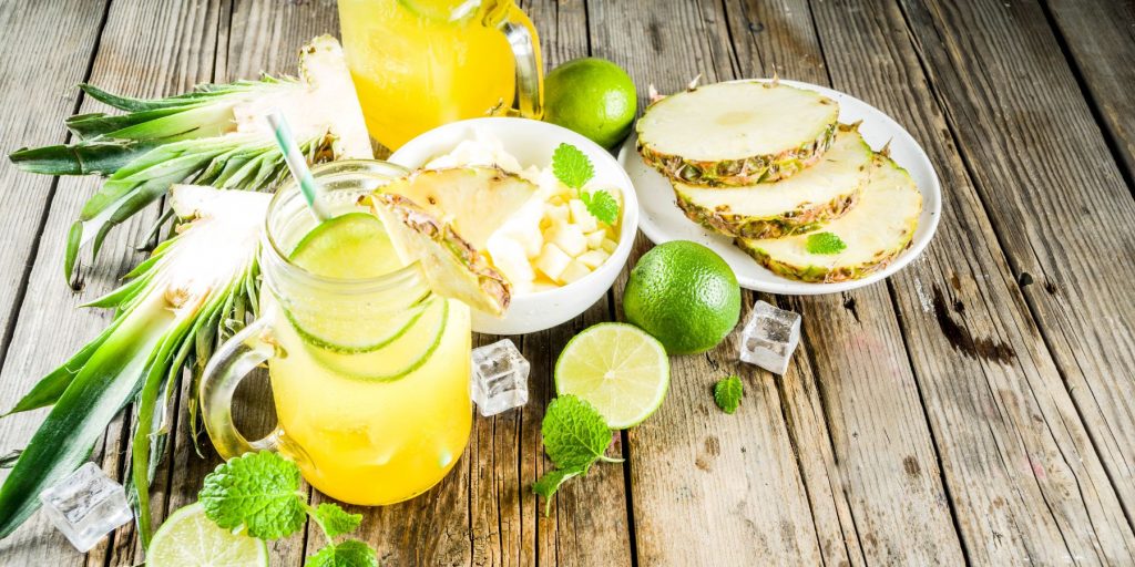 Top-front view of a bright yellow Pineapple Mojito on a wooden surface surrounded by fresh, cut limes, sliced pineapple on a place and mint leaves