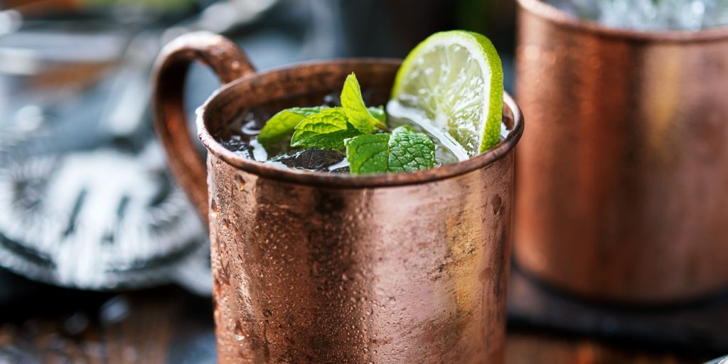 A tight close-up of a pair of Smoky Campfire Moscow Mules in copper mugs on a wooden surface in an outdoor setting