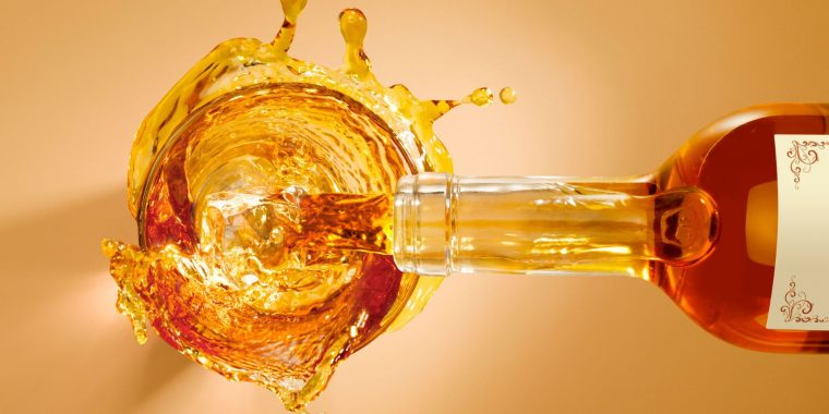 A bottle of amber liquor being decanted into a full glass, making splashes