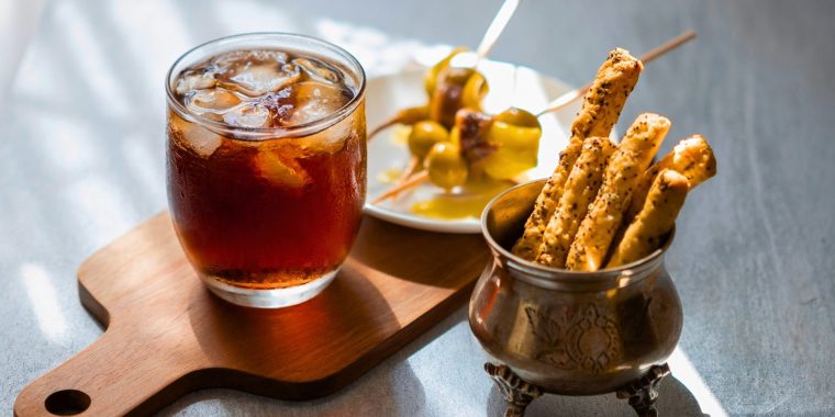 Close up view of a Cynar Spritz cocktail served on a wooden platter with a variety of snacks, presented on a white linen surface