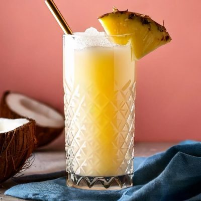 Close-up of a Painkiller cocktail with pineapple garnish, pink background, halved coconuts to the side