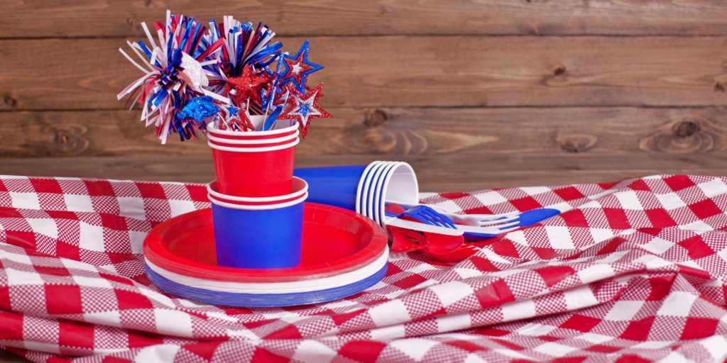 Close up of a red and white tablecloth and collection of red, white and blue paper cups that are ideal to use as decor elements for a Memorial Day party at home