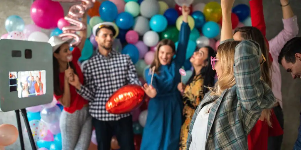 Tight shot of a diverse group of friends taking photos with props against a balloon arch at a photo booth at a Memorial Day party at home