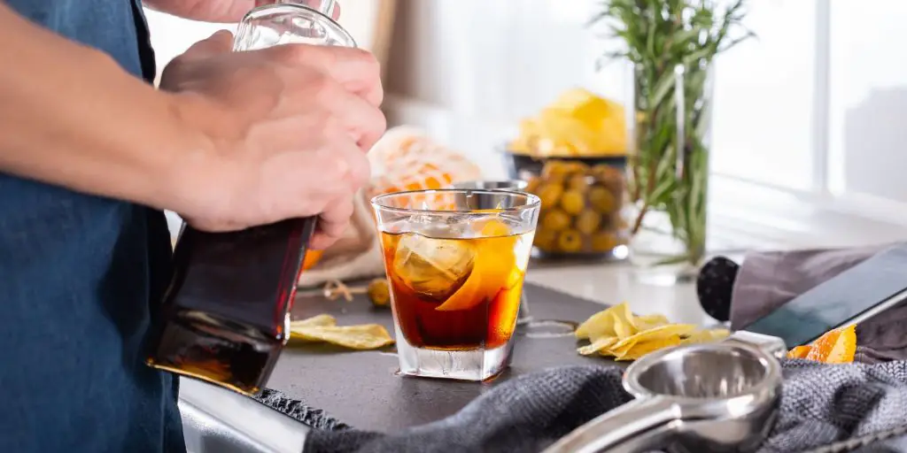 Close up of a home mixologist pouring a Freezer Door Cocktail from a bottle of liquor into a tumbler on a kitchen surface filled with cocktail-making essentials