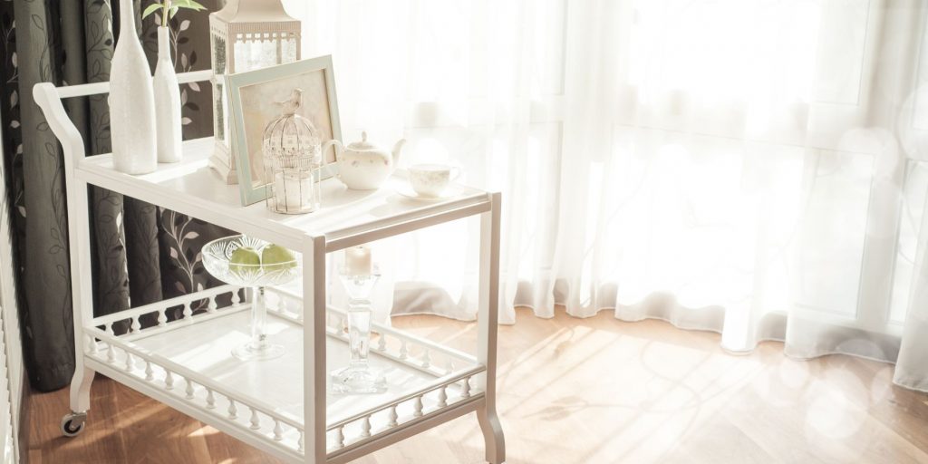 Small white minimalist bar cart in shades of white standing in front of a window in a light-filled room