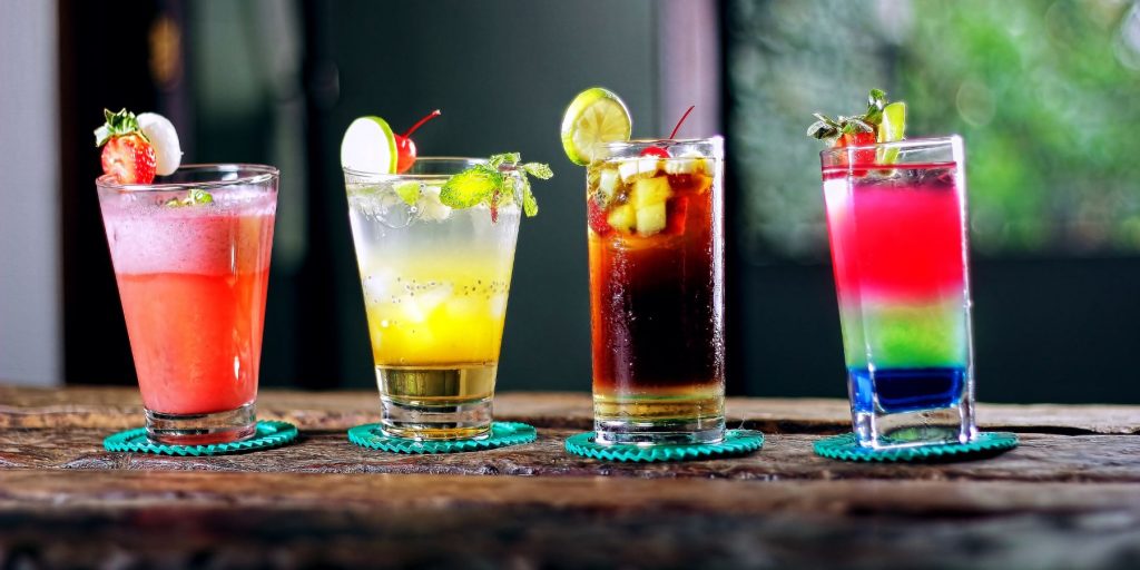Close up of a row of four rainbow colored cocktails set on a wooden surface in an outdoor daytime space