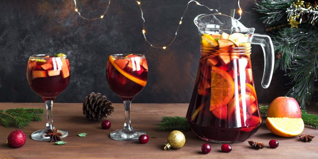 A jug and two glasses of Cranberry Chai Sangria against a festive backdrop with Christmas decorations and strings of fairy lights