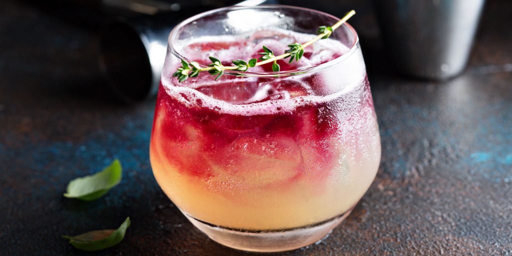 Close up of a Pomegrate Whiskey Sour on a dark table, garnished with a sprig of thyme