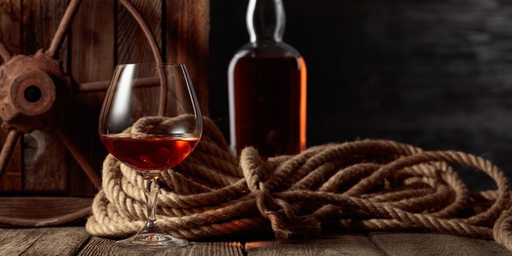 Close up of a snifter of dark rum in a still life set-up with ship's rope, a bottle of dark rum and a spoked wheel in the background