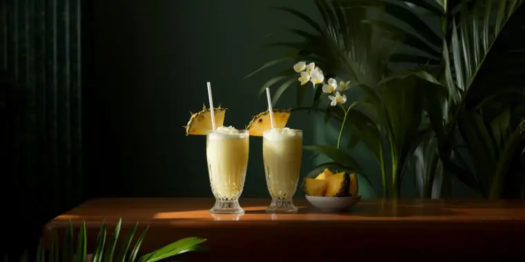 Two Pina Colada cocktails on a table in a dark green room with tropical greenery as decoration