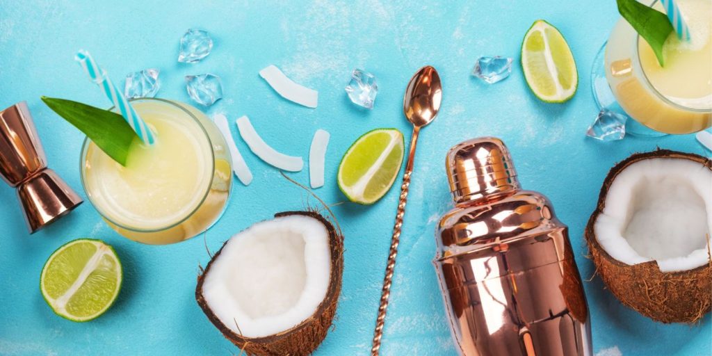 Top view of all the ingredients and tools required to make a Pina Colada cocktail, on a light blue backdrop in a flat lay format