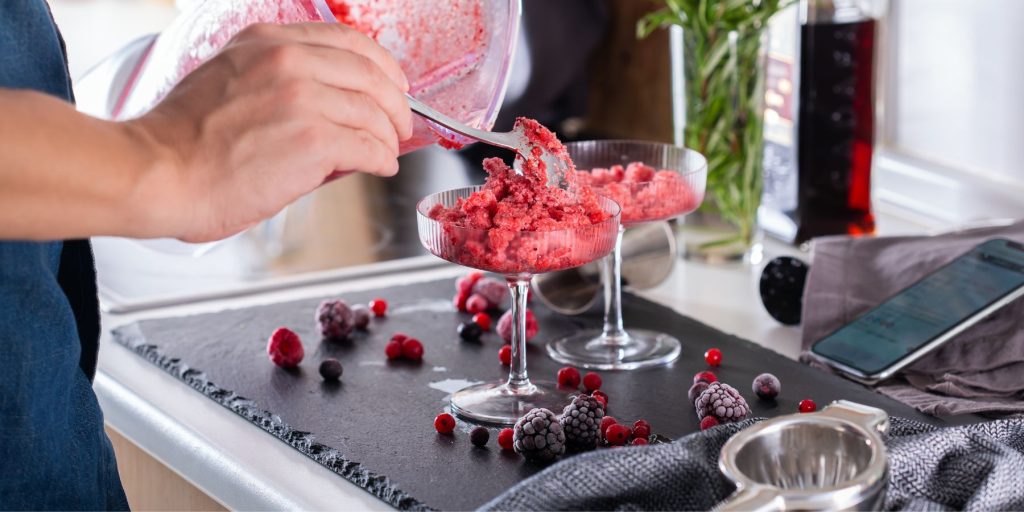 Close up of a home mixologist spooning Frozen Raspberry Daiquiris into serving glasses in a light bright home kitchen
