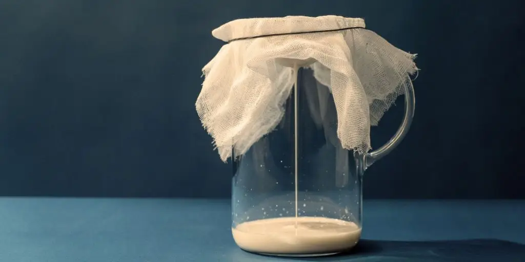 Close up of a jug overlayed with cheesecloth with milk straining into the jug against a blue backdrop