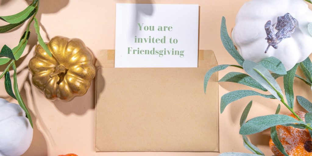 Top view of a Friendsgiving invitation on white paper in a brown paper envelope surrounded by fall inspired decor elements