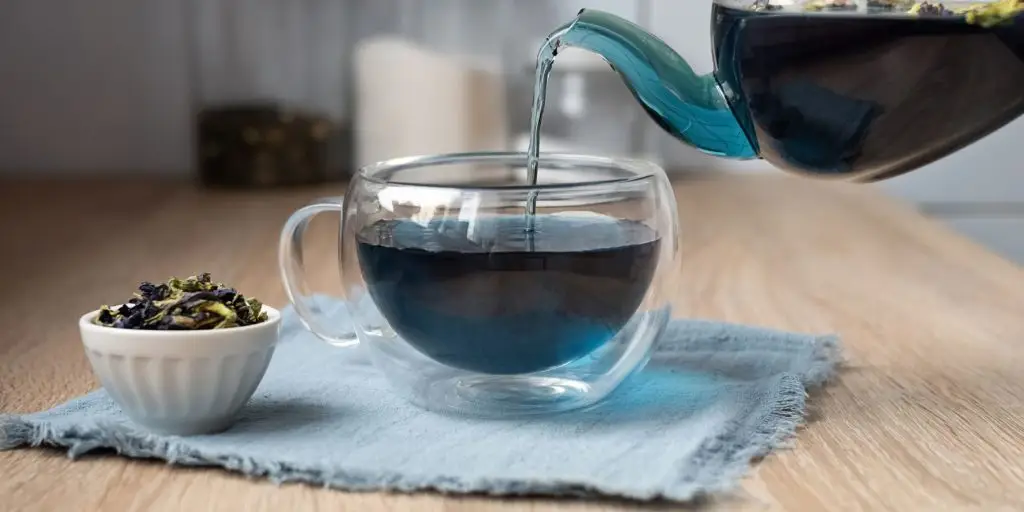 Close up image of a person pouring blue butterfly flower tea from a pot into a clear, perspex cup on a table in a modern kitchen environment 