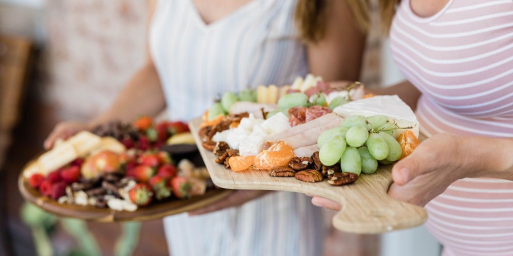Close up image of two women carrying charcuterie platters out to share with their friends at a daytime gathering