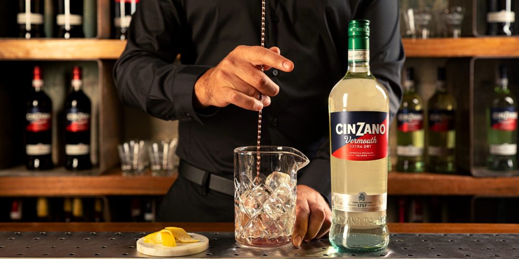A bartender stirring a cocktail in a mixing glass, a bottle of Cinzano Vermouth Extra Dry in the foreground