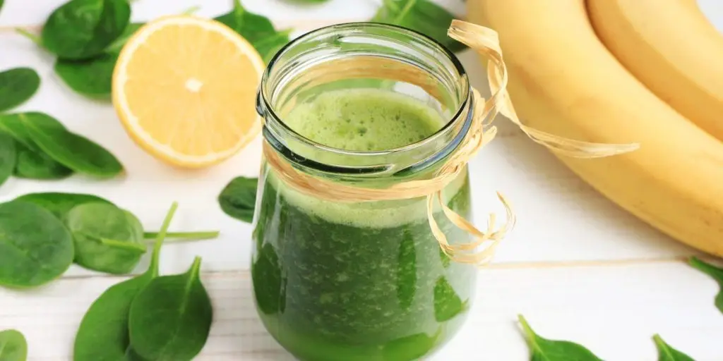 Close up top view of a Green Goddess Smoothie in a mason jar container, surrounded by spinach leaves, sliced lemon and fresh banana