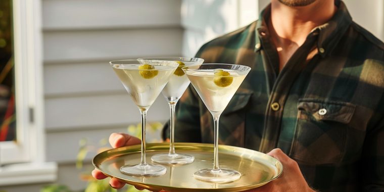 Close up of a man wearing a flannel shirt holding a tray with three Dirty Martini cocktails on it