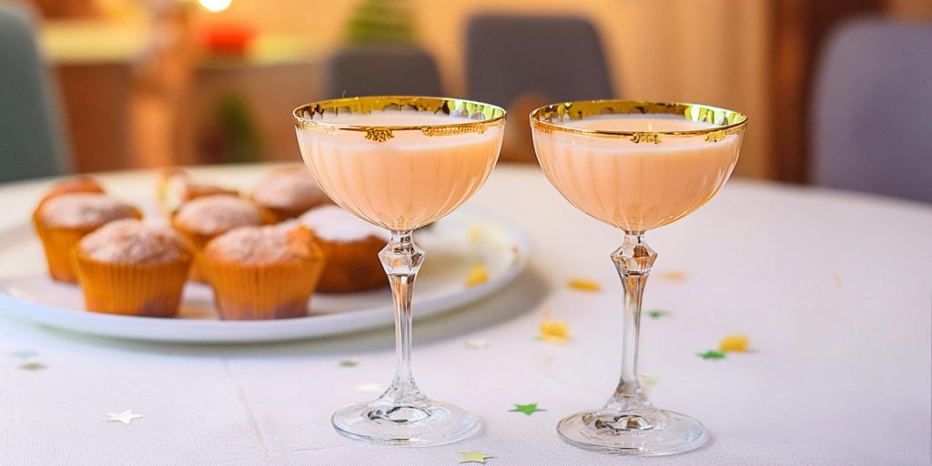 Two Salted Caramel White Russian cocktails in ornate coupe glasses on a table with a white tablecloth next to a serving platter with caramel cupcakes