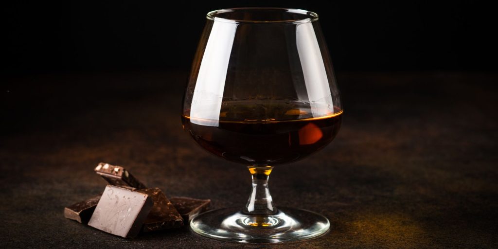 A snifter glass filled with deep amber cognac sits on a dark surface, accompanied by a few pieces of rich, dark chocolate. The scene has a moody, elegant ambiance, highlighting the luxurious pairing of cognac and chocolate.