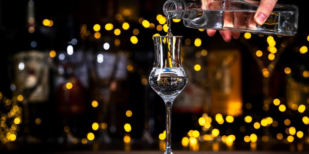 Clear grappa being poured into a traditional narrow glass with golden bokeh lights in the background, photo by 5ph/Adobe Stock