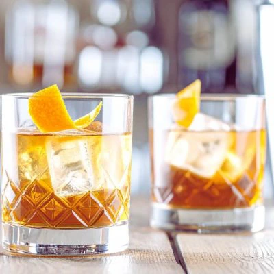 Two Whiskey Old Fashioned cocktails on a bar counter, bottles of bourbon in the background