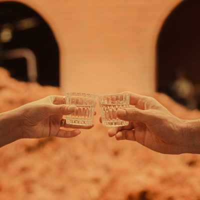 Two people clink together shot glasses of tequila in front of a tequila-making location in Mexico.
