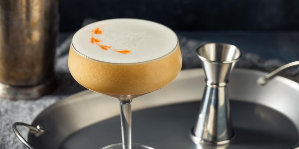 Alt text: Beretta’s Rattlesnake cocktail in a coupe glass with a creamy foam and bitters garnish, accompanied by maple leaves on a rustic surface. Image by Brent Hofacker on Adobe Stock.