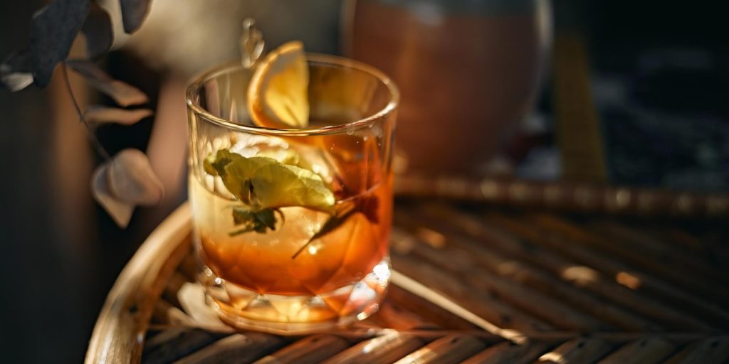 Expense Account cocktail in a short glass with citrus garnish, bathed in warm light on a wooden table, with maple leaves in the background. Image by phanuphongthepnin on Canva.
