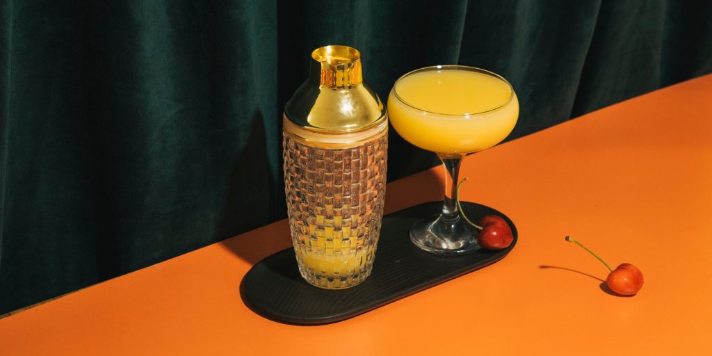 Maple Brown Derby, a maple syrup cocktail served in a coupe glass with a golden shaker beside it, set on an orange surface with two cherries. Image by Studio Germany on Canva.